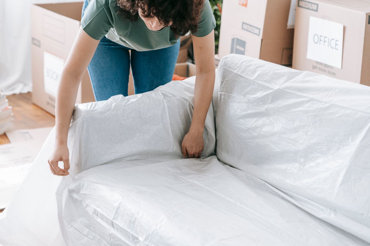 why-choose-us-02 A woman carefully covers a sofa with protective wrapping while packing boxes in a bright room.