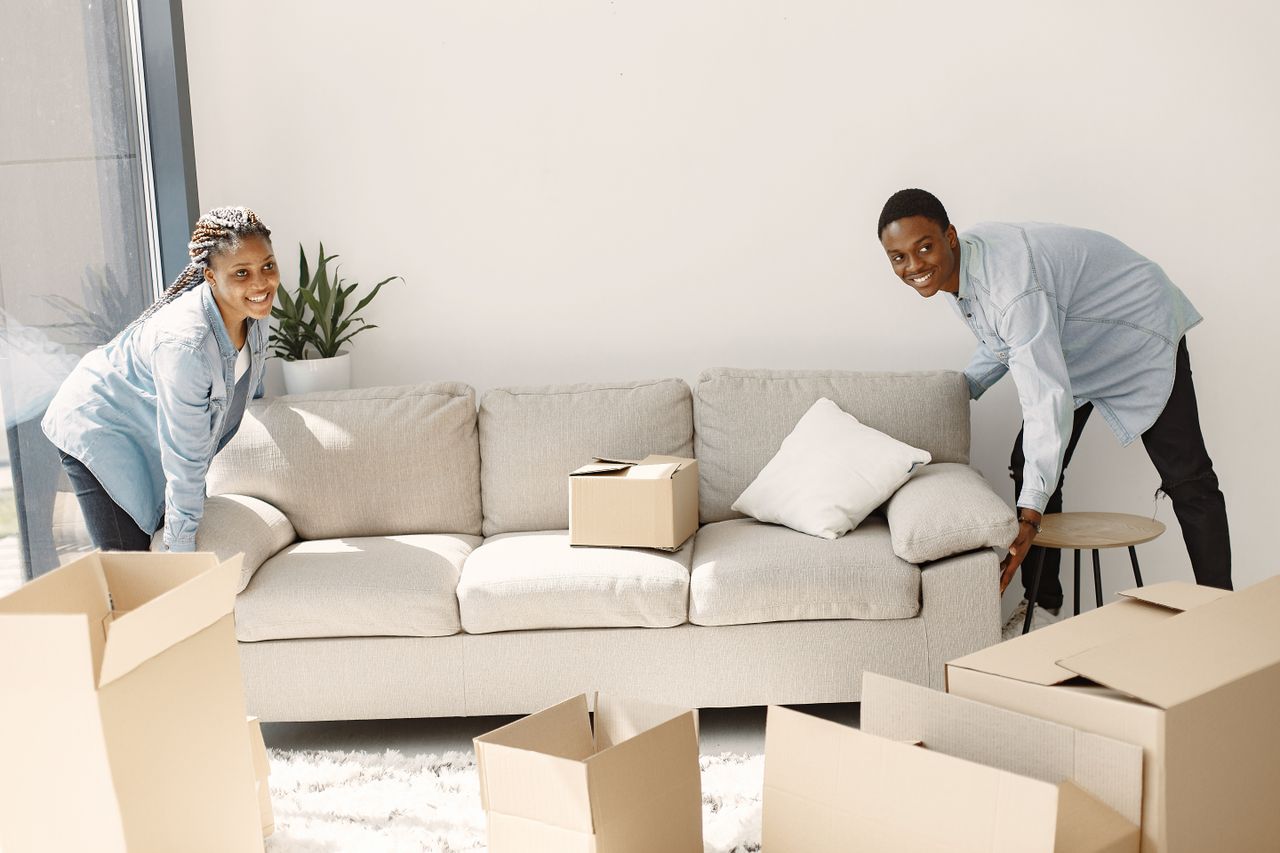 our-value A cheerful couple moving furniture in a well-lit modern living room with cardboard boxes.