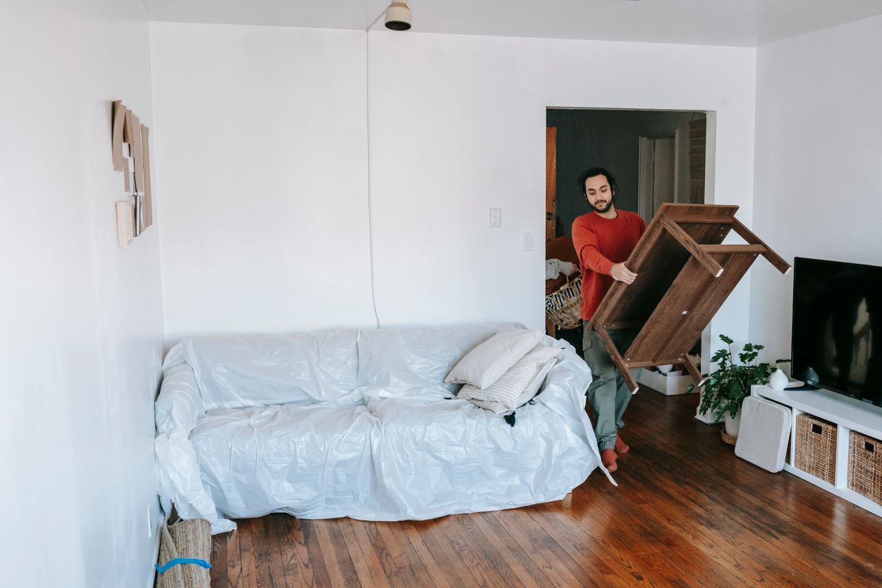 services-01 A man is carrying a wooden table in a bright living room beside a wrapped sofa, getting ready for a move.